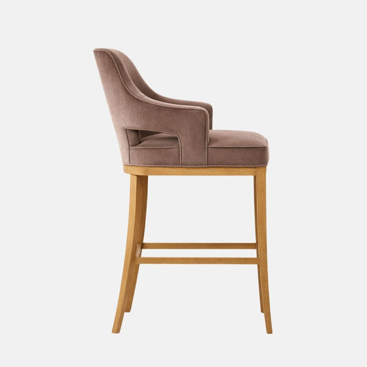 Side view of a taupe/grey velvet bar-height stool with a curved wraparound back and arms, open arm cutouts, thick seat cushion, and light oak legs with stretchers, on a white background.