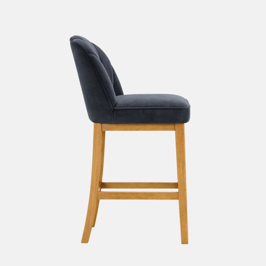 Side view of a dark navy upholstered bar stool with a gently curved back and padded seat, set on light oak wooden legs with a horizontal footrest rail, photographed on a white background.