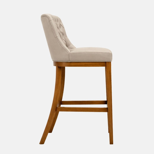 Side view of a beige upholstered bar stool with a tufted backrest, smooth padded seat, and tapered wooden legs with lower stretcher rails.