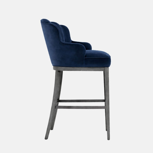 Side view of a bar-height stool upholstered in navy blue velvet with a wraparound curved back and integrated arms, a thick cushioned seat, and a grey wooden frame with four legs and stretchers, photographed on a white background.