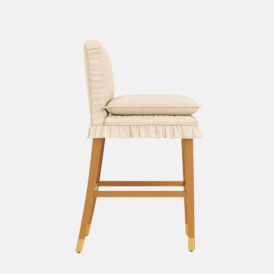 Side view of a cream upholstered bar stool with a padded back, loose seat cushion, ruffled skirted base, wooden legs with brass caps, and a wooden footrest, on a white background.