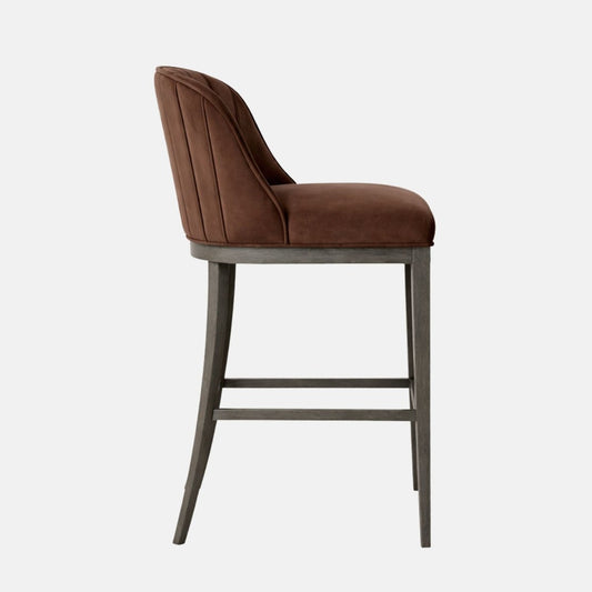Side view of a bar-height stool upholstered in brown suede/velvet with a curved, channel-stitched backrest and a thick cushioned seat, on a dark grey wooden four-leg frame with two horizontal stretcher bars, photographed on a white background.