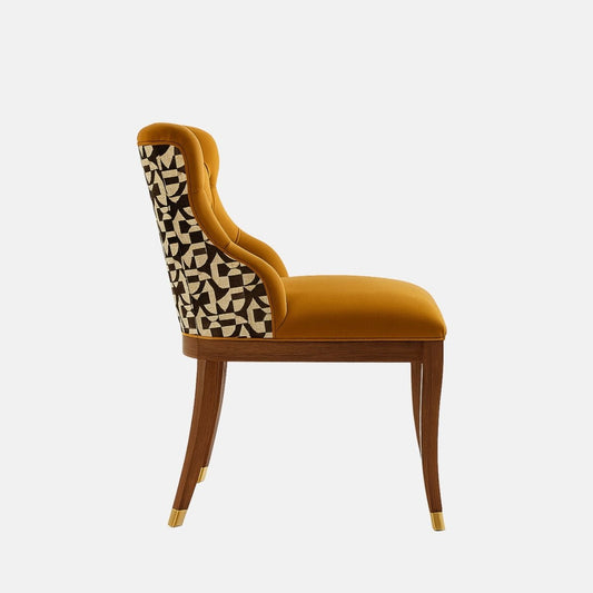 Upholstered accent chair in mustard-gold velvet with a button-tufted backrest, curved silhouette, patterned fabric on the outer back, and dark wooden legs with brass-capped feet, shown on a white background
