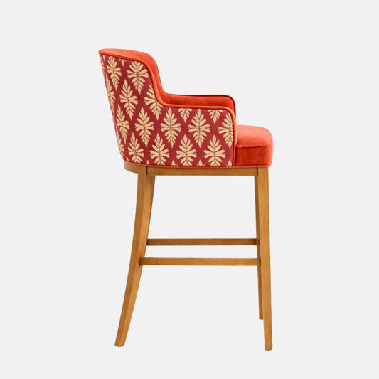 Side view of an orange upholstered bar stool with a curved wraparound back and integrated arms, a red patterned outer back panel with cream leaf motifs, thick cushioned seat, and a light oak wooden frame with square stretchers, photographed on a white background.