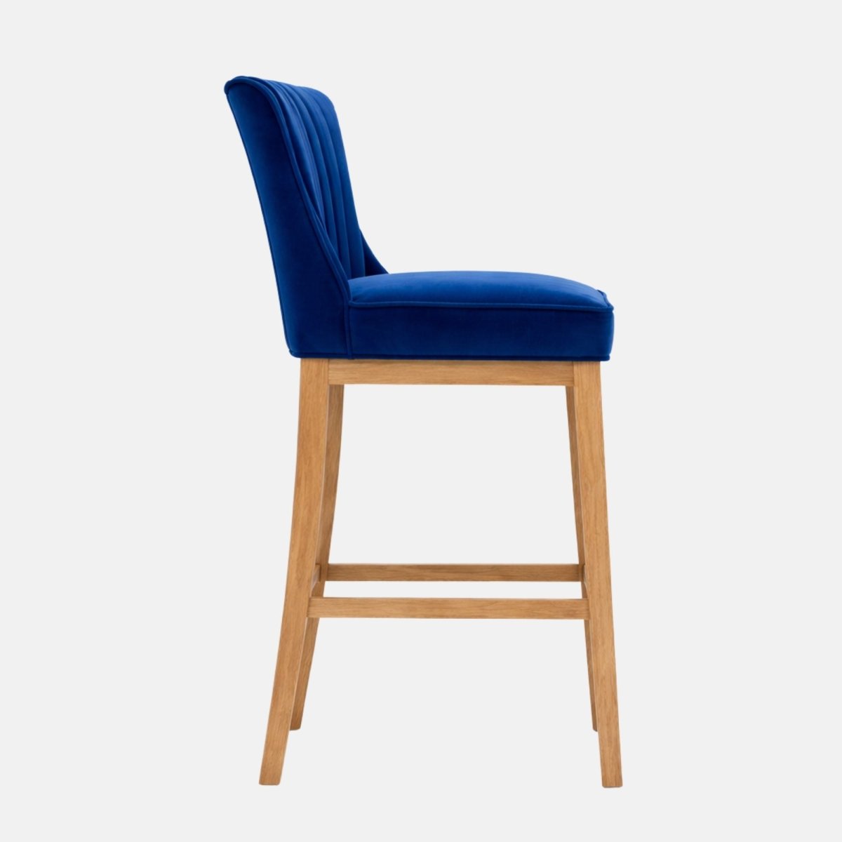 Side view of a royal blue velvet bar stool with a channel-tufted backrest, padded seat, and light wood legs with lower stretcher rails.