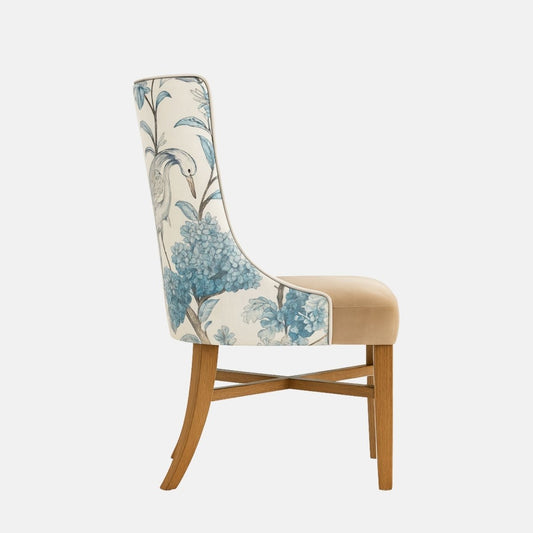 Upholstered dining chair with a beige cushioned seat and a high, gently curved backrest upholstered in a blue botanical and bird-patterned fabric, supported by natural wooden legs, shown on a white background