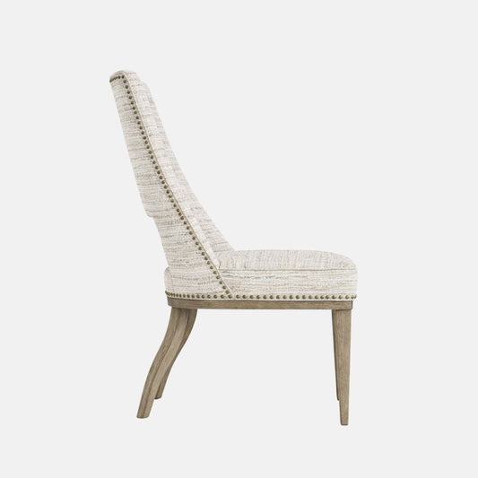 Upholstered dining chair in light beige textured fabric with a high back featuring a rectangular cut-out detail, nailhead trim, cushioned seat, and light wooden legs, shown on a white background