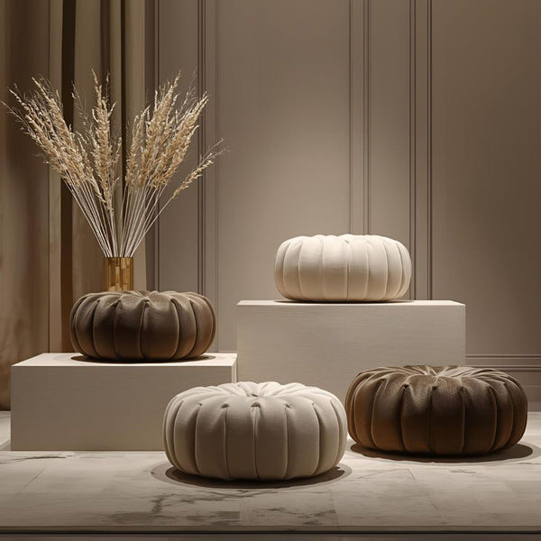 Four ottomans are lined up against a beige backdrop. Designs include: cream hexagon with gold base, geometric brown/taupe with fringe, golden velvet checkerboard with smooth base, and off-white with gold line patterns on a dark wood base.