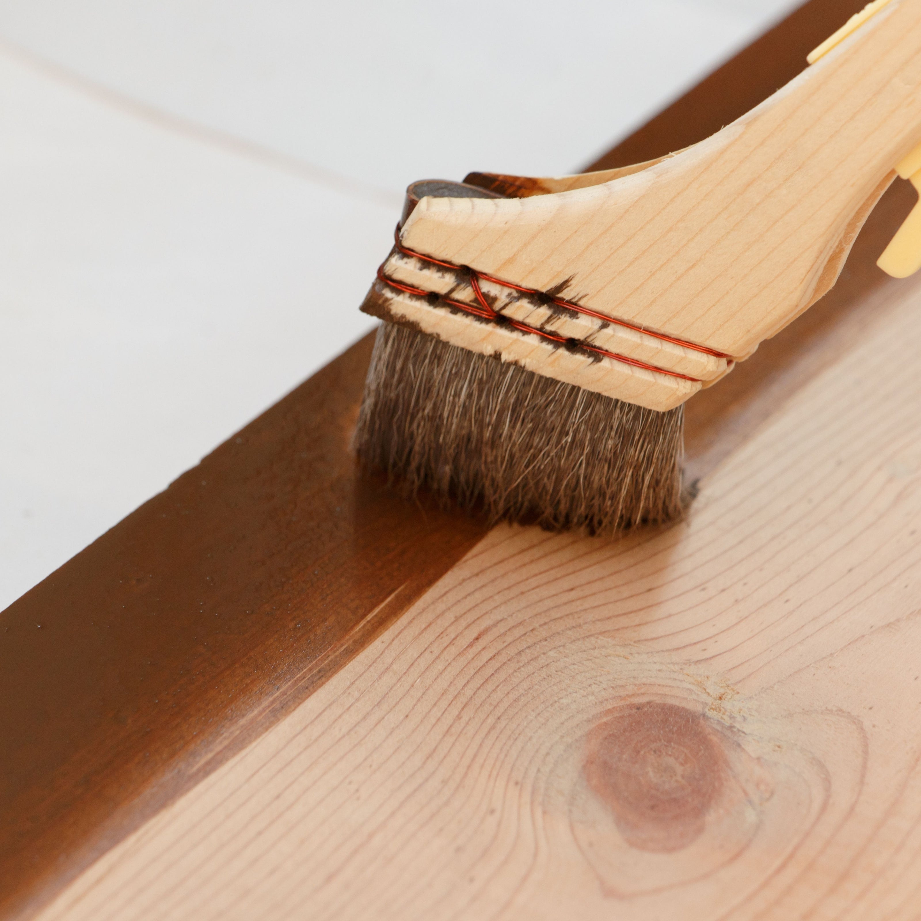 Close-up of a brush applying wood stain or finish to a natural wooden surface, highlighting the grain and craftsmanship of custom furniture making.