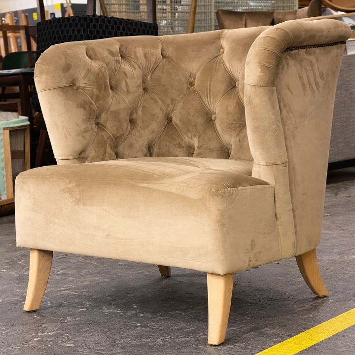 A beige button-tufted upholstered tub chair with curved arms, a plush seat, and light wooden legs, placed in a furniture workshop with other seating in the background.
