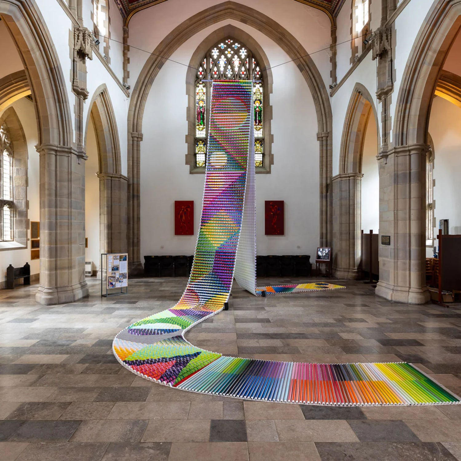 Large-scale contemporary art installation of a colourful woven textile flowing across the stone floor and hanging from the ceiling inside a historic church with arched windows and stained glass.