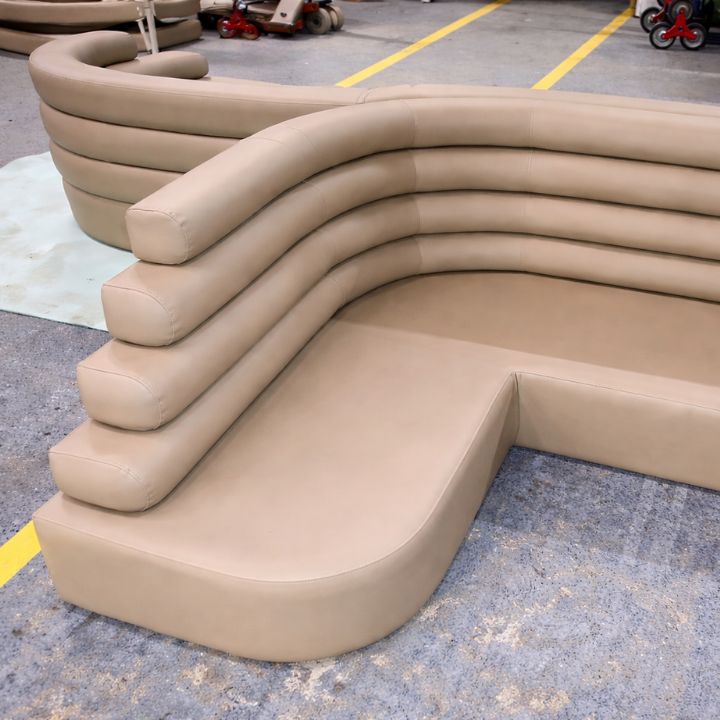 A beige curved modular sofa with a layered, tiered backrest and smooth upholstered finish, arranged in a furniture workshop with equipment and materials in the background