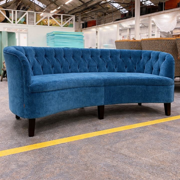 A blue curved upholstered sofa with button-tufted backrest and dark wooden legs, positioned in a furniture workshop with stacked foam and other seating in the background.
