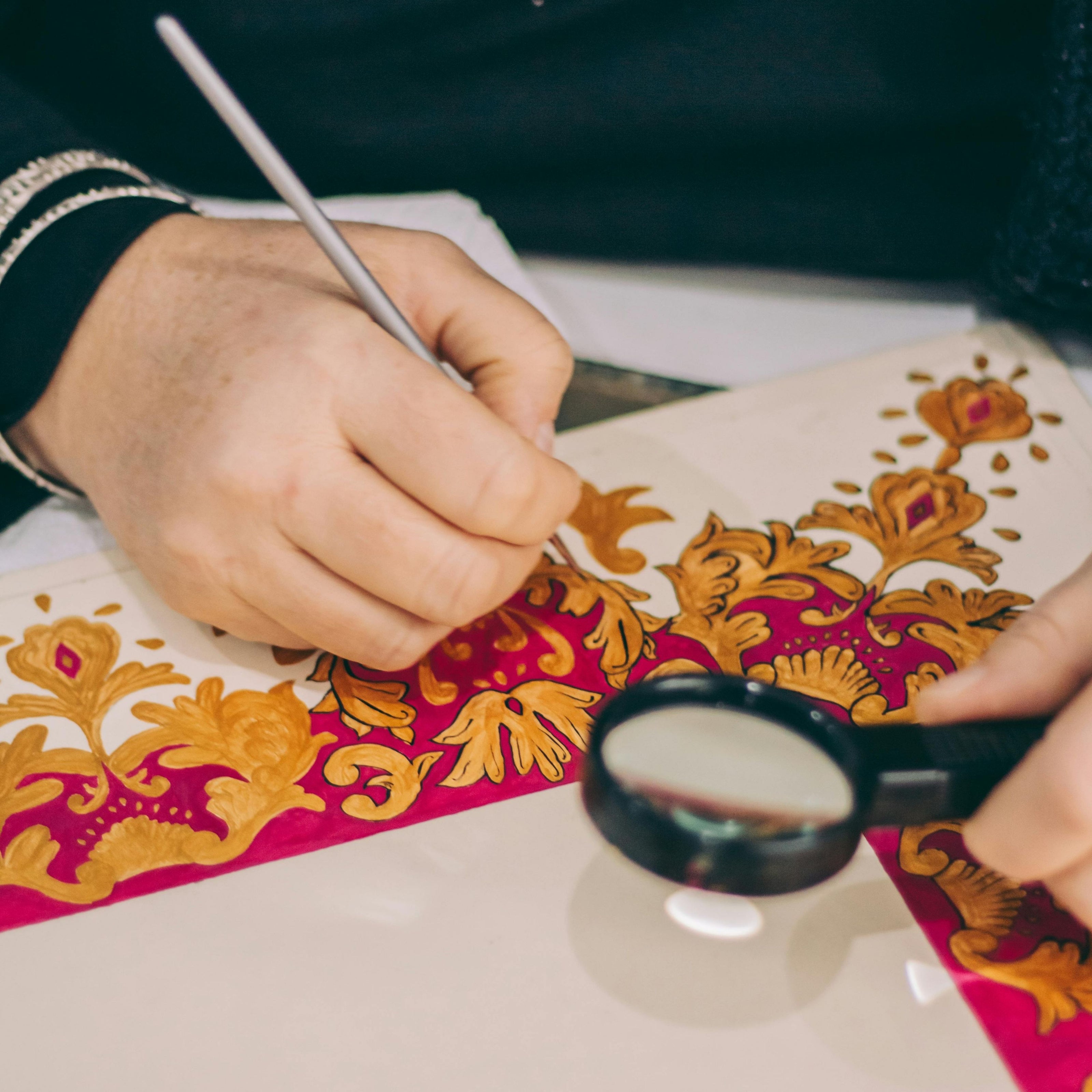 Close-up of an artisan hand-painting an intricate gold and red floral pattern on fabric, using a fine brush and magnifying glass for detailed textile design.