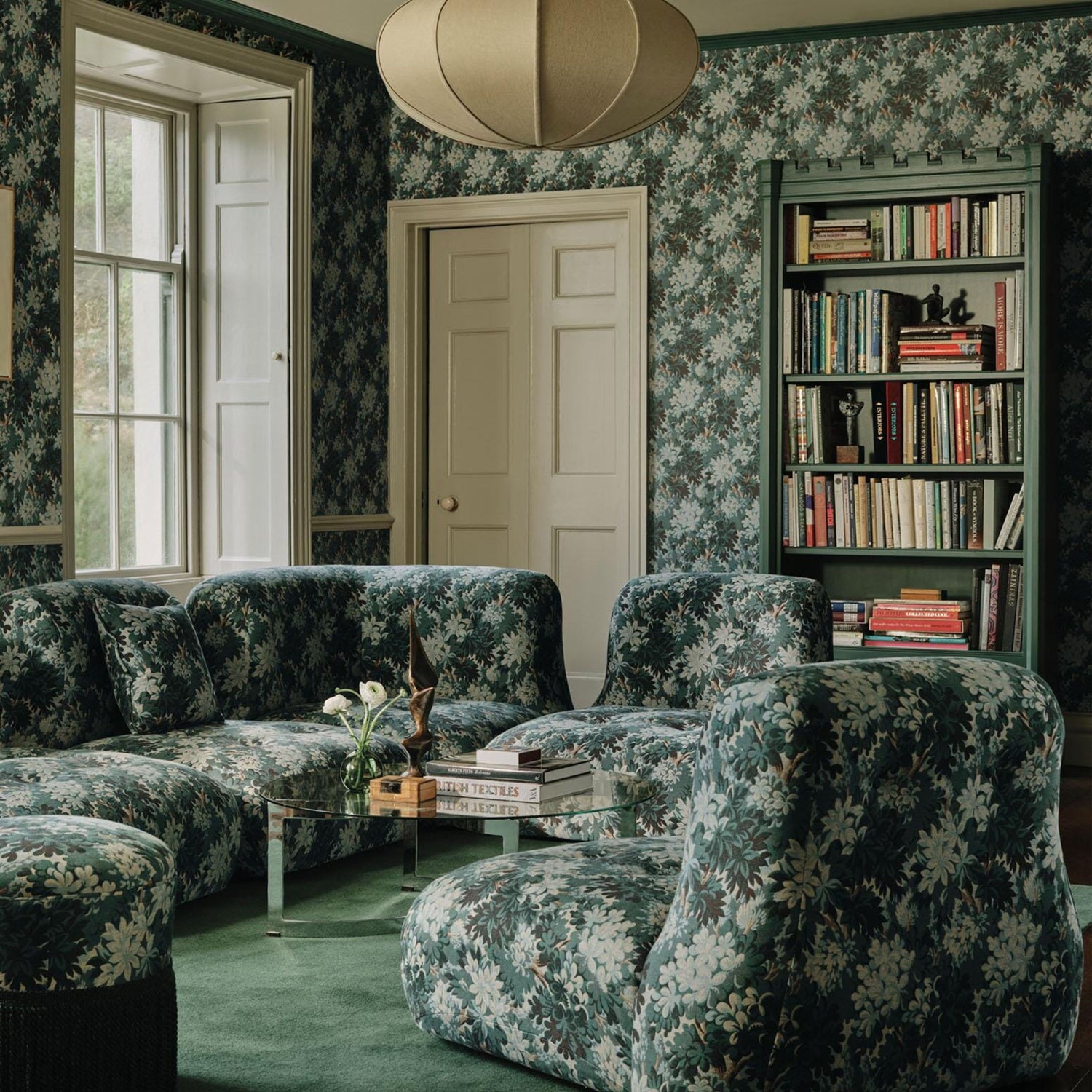Elegant living room with green floral wallpaper, matching floral upholstered sectional sofa, green carpet, and a built-in bookcase filled with books