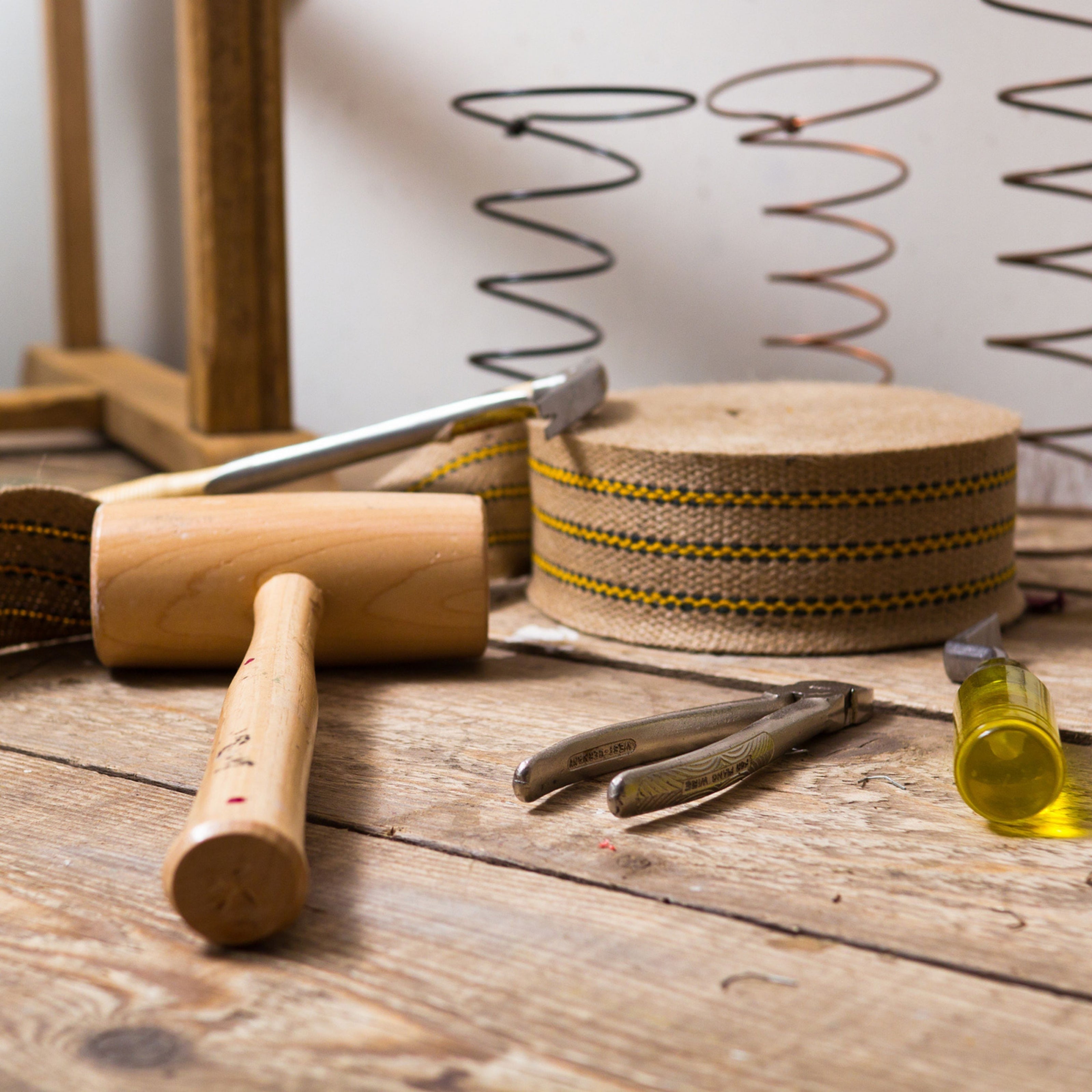 Traditional upholstery tools and materials on a wooden workbench, including a wooden mallet, webbing roll, metal springs, pliers, and tack hammer for handcrafted furniture making.