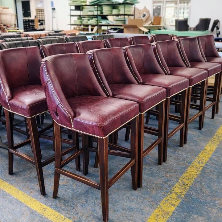 A row of burgundy leather upholstered bar stools with studded trim and wooden legs, arranged in a furniture workshop with materials and equipment in the background.