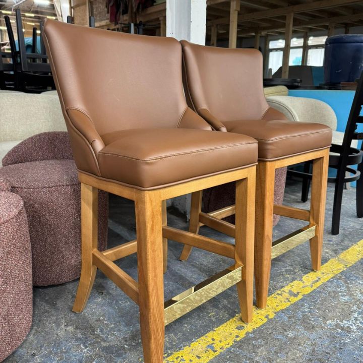 Modern bar stool with brown leather upholstered seat and backrest, featuring wooden legs and brass footrest, ideal for bars and hospitality interiors