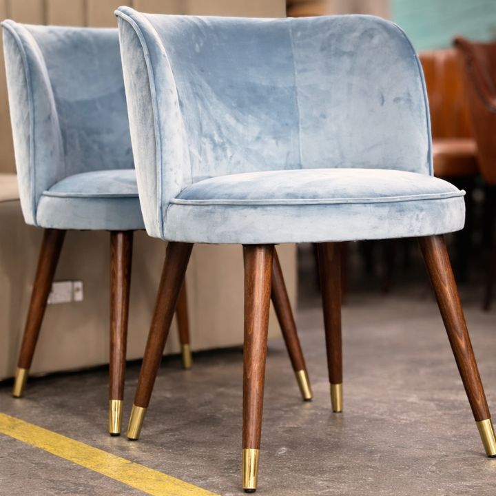 Light blue velvet dining chair with curved backrest and wooden legs with gold tips, ideal for restaurant and modern interiors