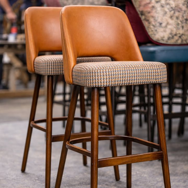 Two upholstered bar stools with tan leather backs, patterned fabric seats, and wooden legs with footrests, placed in a furniture workshop with other seating in the background.