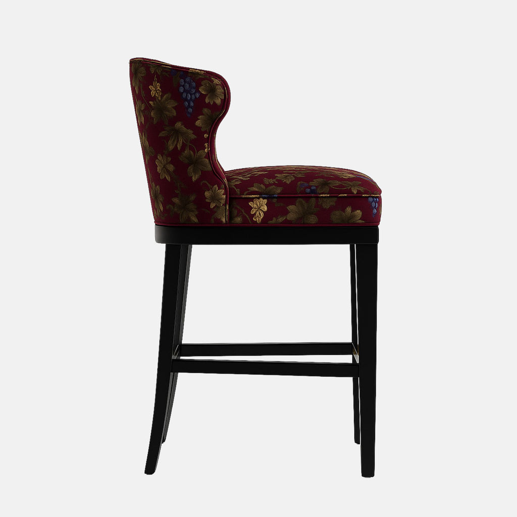 Elegant bar stool with a curved backrest and floral-patterned upholstery in rich burgundy fabric featuring green and purple tones. The frame is black wood with a brass footrest accent, blending classic and contemporary design elements.