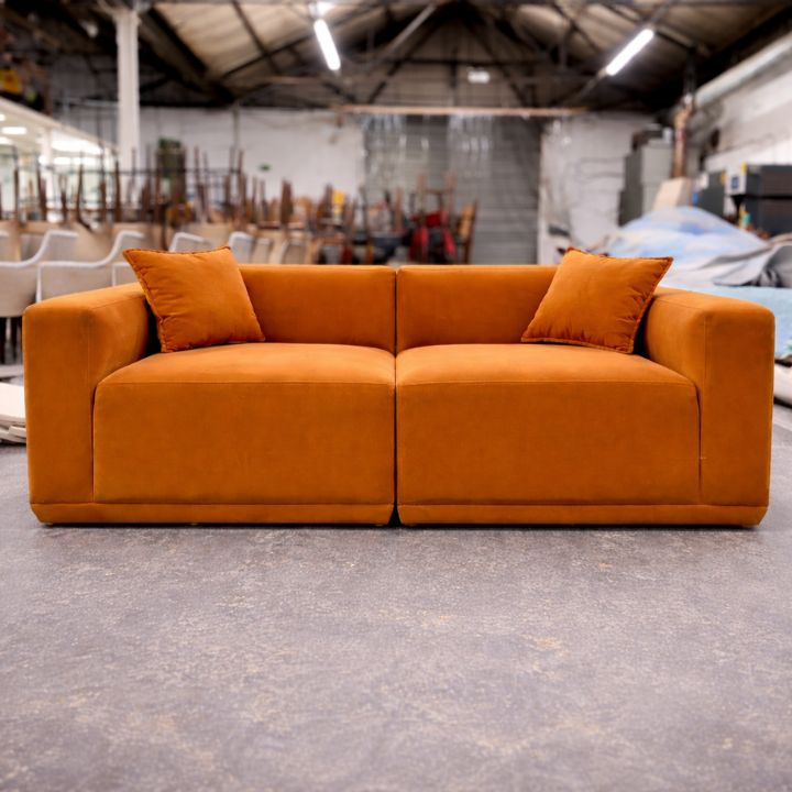 A low-profile orange upholstered sofa with two seat cushions and matching scatter cushions, placed in a spacious furniture workshop with stacked chairs and materials in the background.