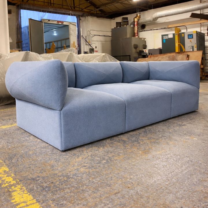 A light blue modular upholstered sofa with rounded arms and deep cushioned seats, placed in a furniture workshop with covered items and equipment in the background.