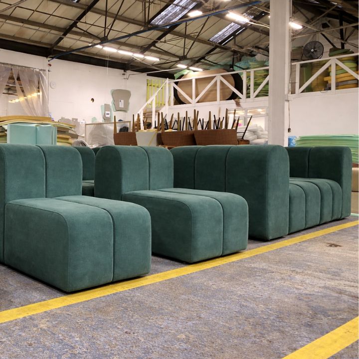 A row of green upholstered modular lounge chairs with rounded edges and low backs, arranged in a furniture workshop with foam stacks and equipment in the background