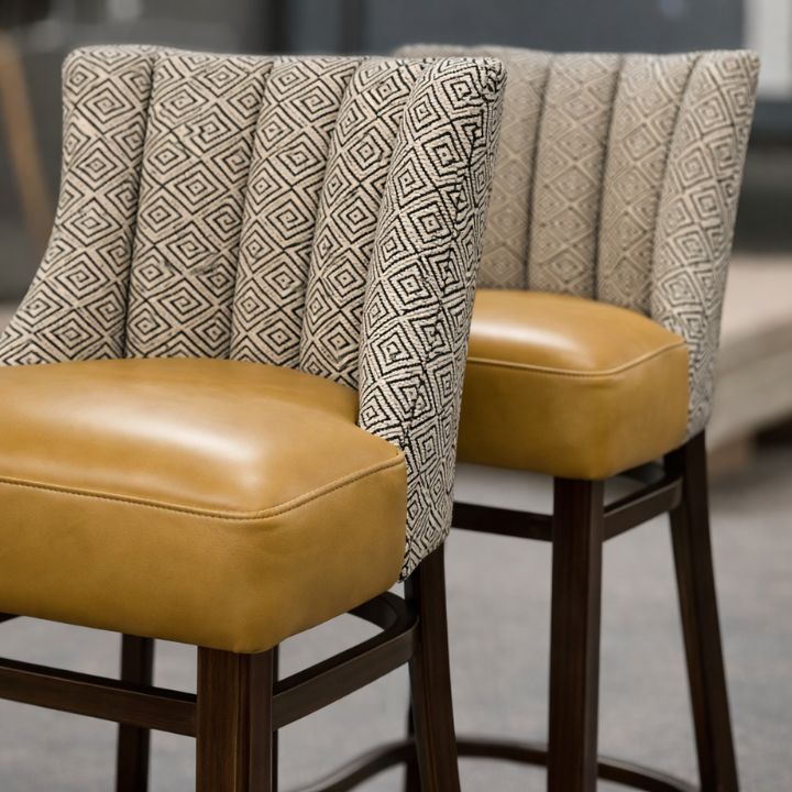 Modern bar stool with geometric patterned fabric backrest and tan leather seat, featuring wooden legs, ideal for bars and hospitality interiors