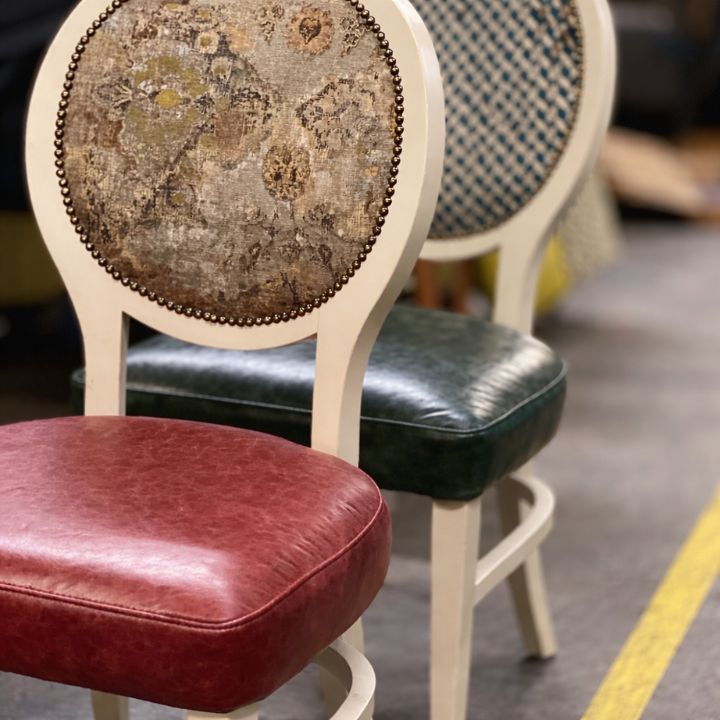 Dining chairs with round upholstered backs featuring patterned fabric and nailhead trim, paired with coloured leather seats and white wooden frames in a showroom.