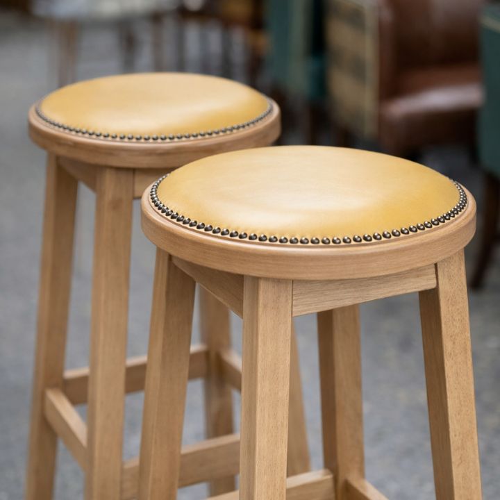 Wooden bar stool with round leather seat and decorative stud trim, featuring a natural wood frame, ideal for bars and hospitality interiors