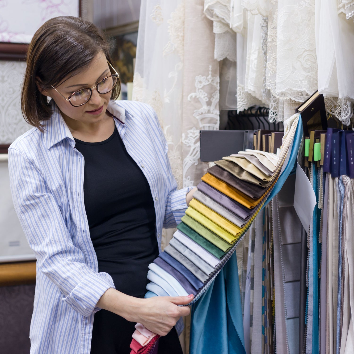 Woman wearing glasses and a striped shirt examining a sample book of colourful fabric swatches in a textile or fabric store.