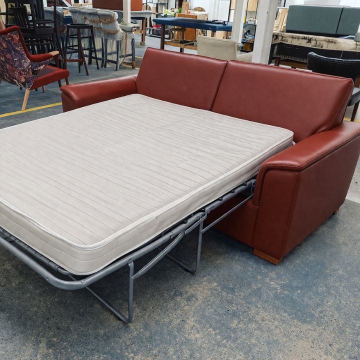 A red upholstered sofa bed extended into a bed, featuring a metal folding frame and a beige mattress, set inside a furniture workshop with chairs and materials in the background.