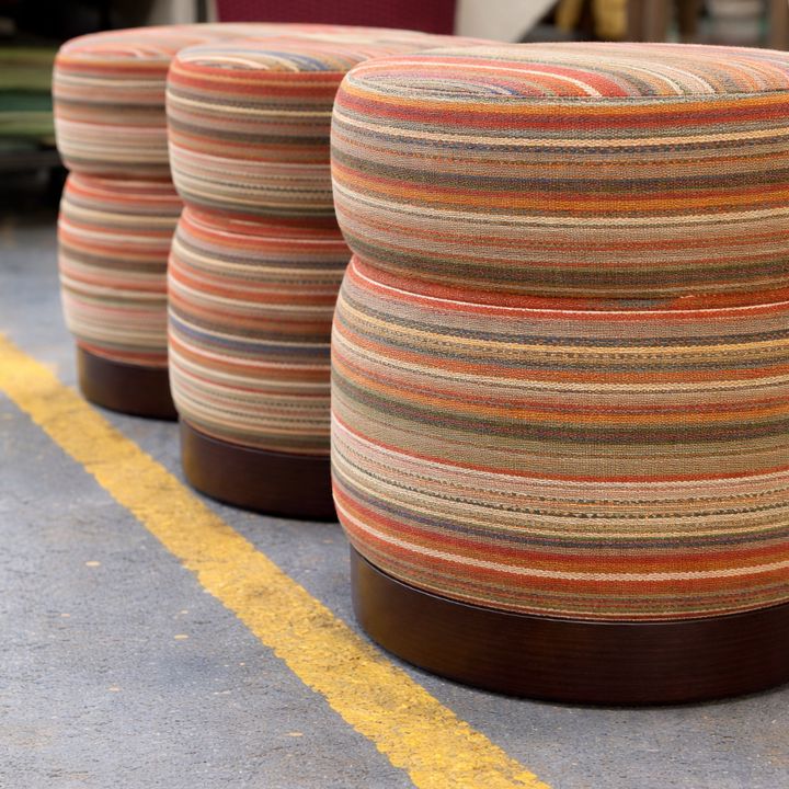 Round upholstered pouffe with multicolour striped fabric and wooden base, ideal for lounges, hotels, and modern interiors
