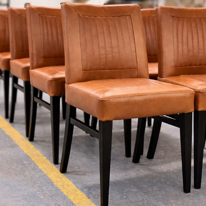 Tan leather dining chairs with vertical stitched backs and black wooden legs arranged in rows in a showroom.