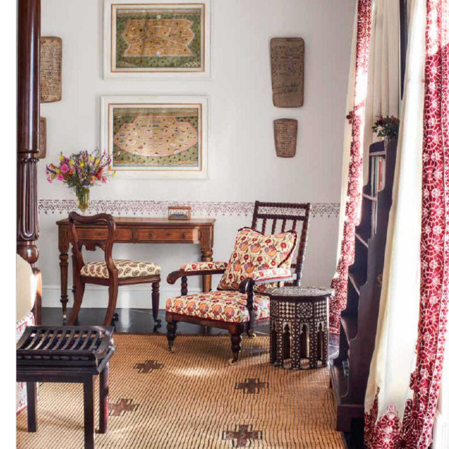 Traditional room with antique wooden desk and chairs upholstered in red and cream patterned fabric, woven rug with cross motifs, and red patterned curtains