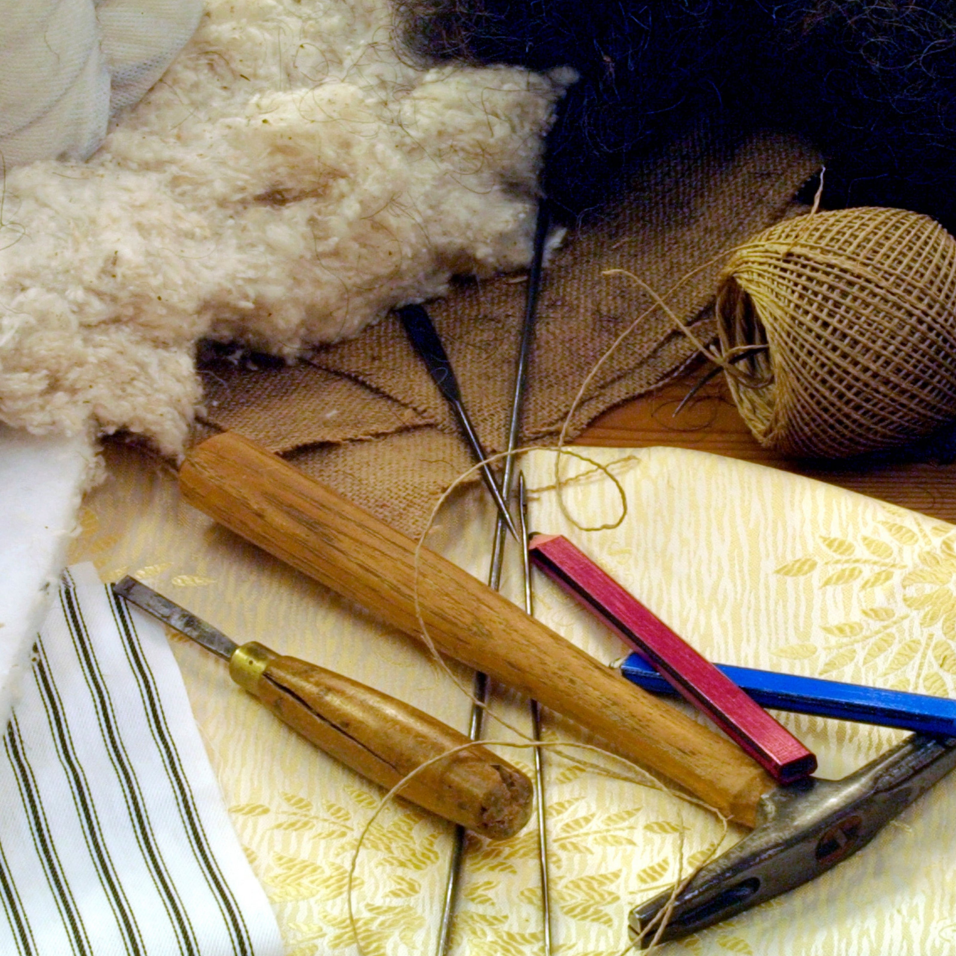 Close-up of traditional upholstery tools and materials, including natural fibers, twine, fabric samples, chisels, and hammers used for handcrafted furniture making.