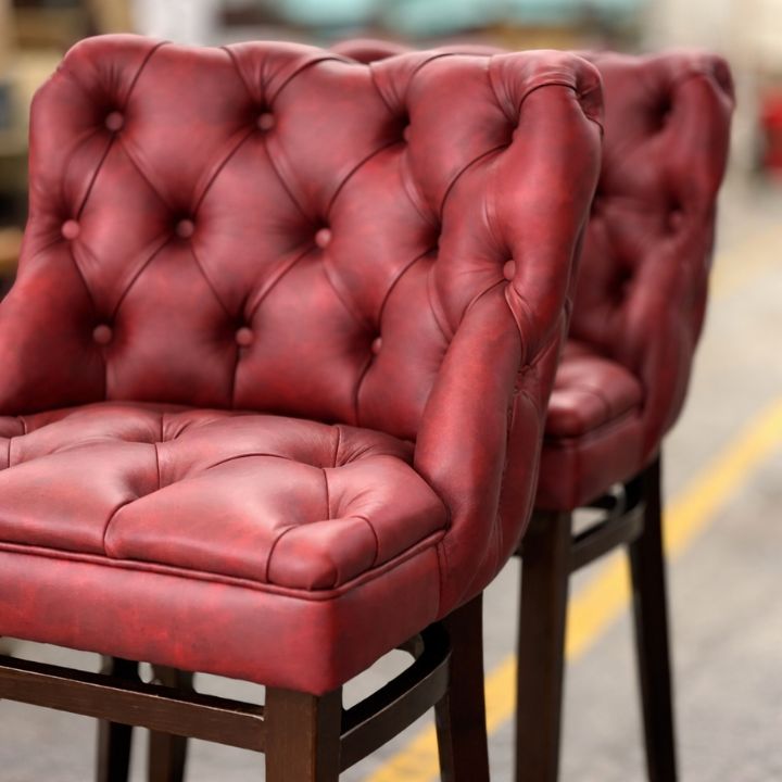 Red button-tufted leather bar stools with arms and wooden frames arranged in a showroom.