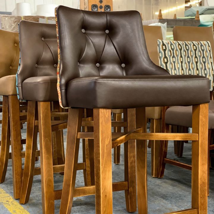 Brown button-tufted leather bar stools with wooden legs and footrests displayed in a furniture showroom.