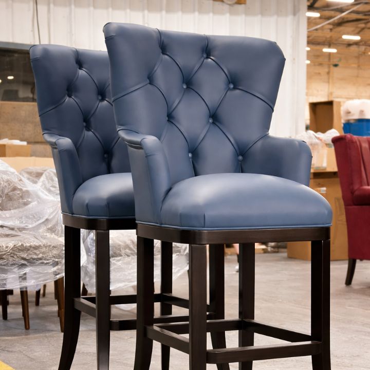 Blue button-tufted upholstered bar stools with arms and dark wooden frames displayed in a warehouse showroom.
