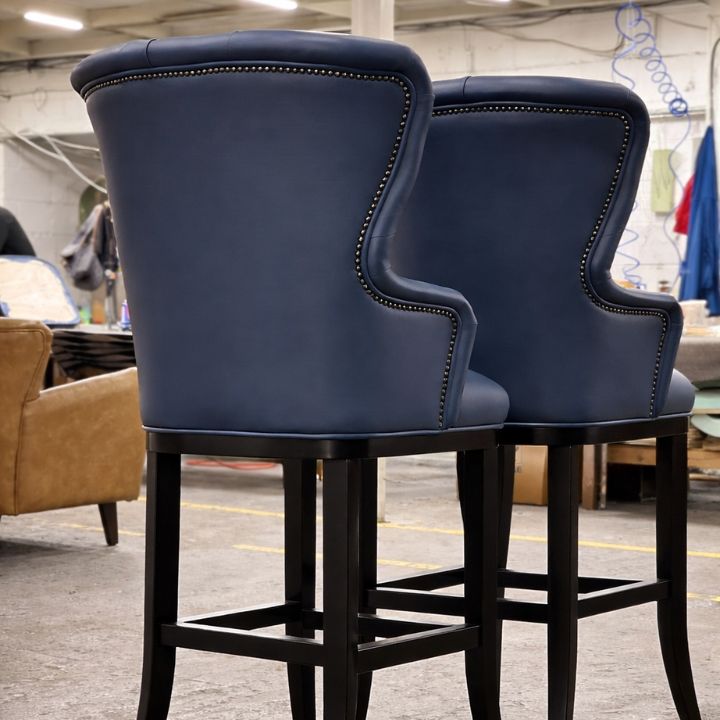 Two blue upholstered high-back bar chairs with curved backs, studded trim detailing, and dark wooden legs, positioned in a furniture workshop with other seating and materials in the background.