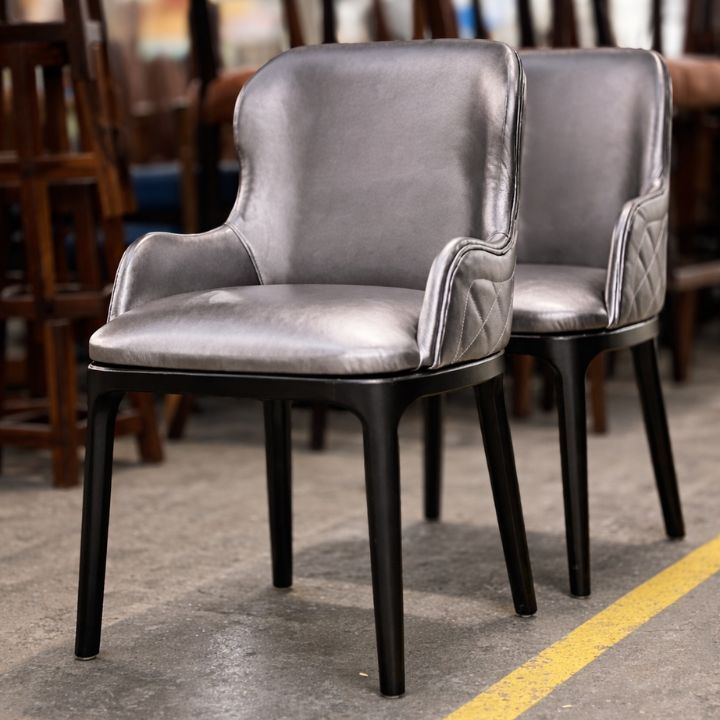 Two grey upholstered dining chairs with curved arms, quilted side detailing, and black wooden legs, placed in a furniture workshop with other chairs in the background.