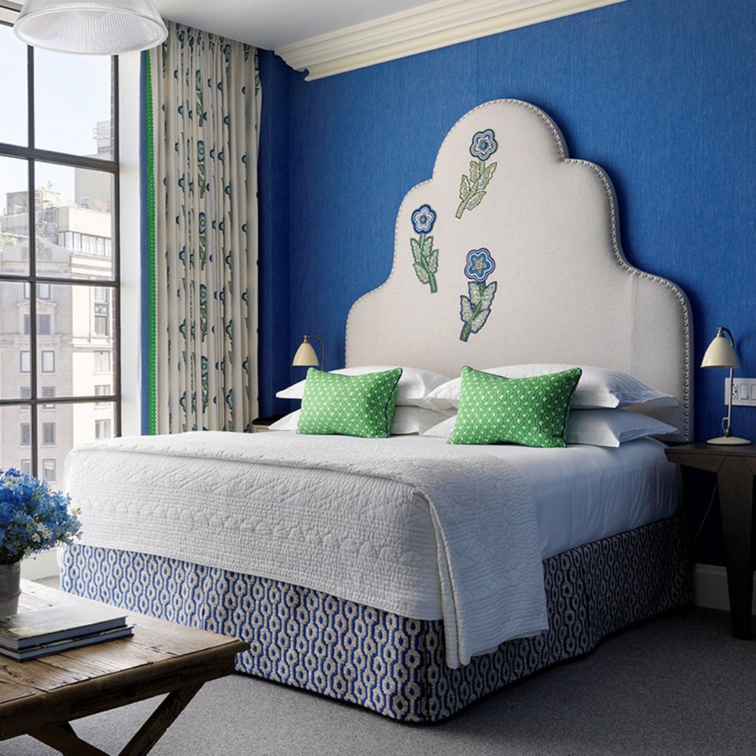 Bright blue and white bedroom with scalloped upholstered headboard decorated with embroidered flowers, patterned bed skirt, and green accent pillows.