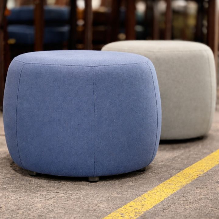 Two rounded upholstered ottomans, one in blue and one in light grey fabric, placed on a workshop floor with chairs in the background.