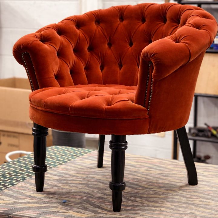 A red button-tufted upholstered tub chair with curved arms, stud detailing, and dark wooden legs, placed on a workbench in a furniture workshop.