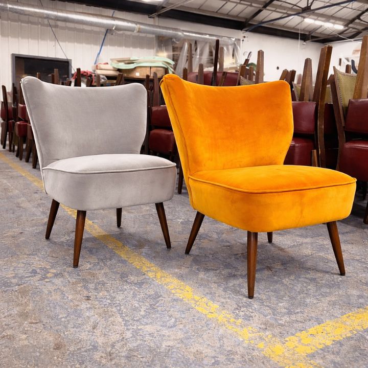 Two upholstered accent chairs, one in light grey and one in bright orange velvet, with curved backs and wooden legs, placed in a furniture workshop with stacked chairs in the background.
