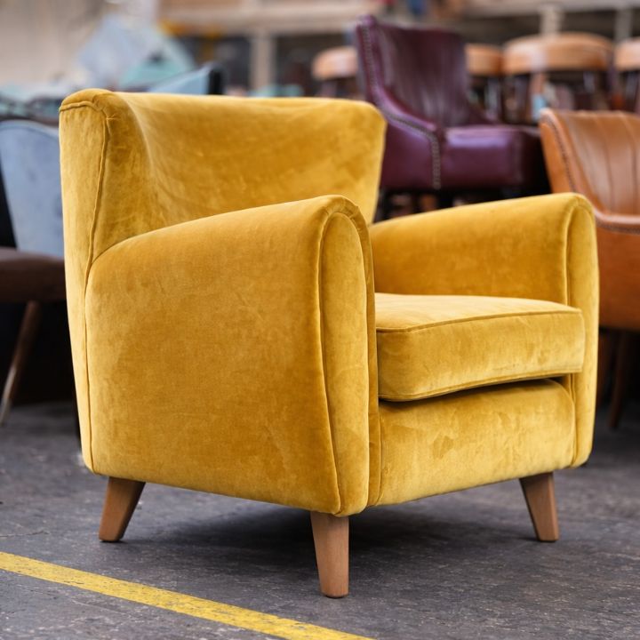 A mustard yellow velvet upholstered armchair with rounded arms and wooden legs, placed in a furniture workshop with other chairs blurred in the background.