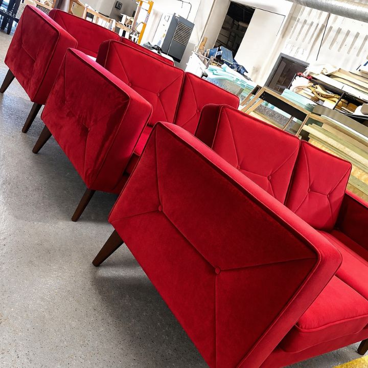 A row of red upholstered armchairs with angular arms, button detailing, and wooden legs, arranged inside a furniture workshop with materials and equipment in the background.
