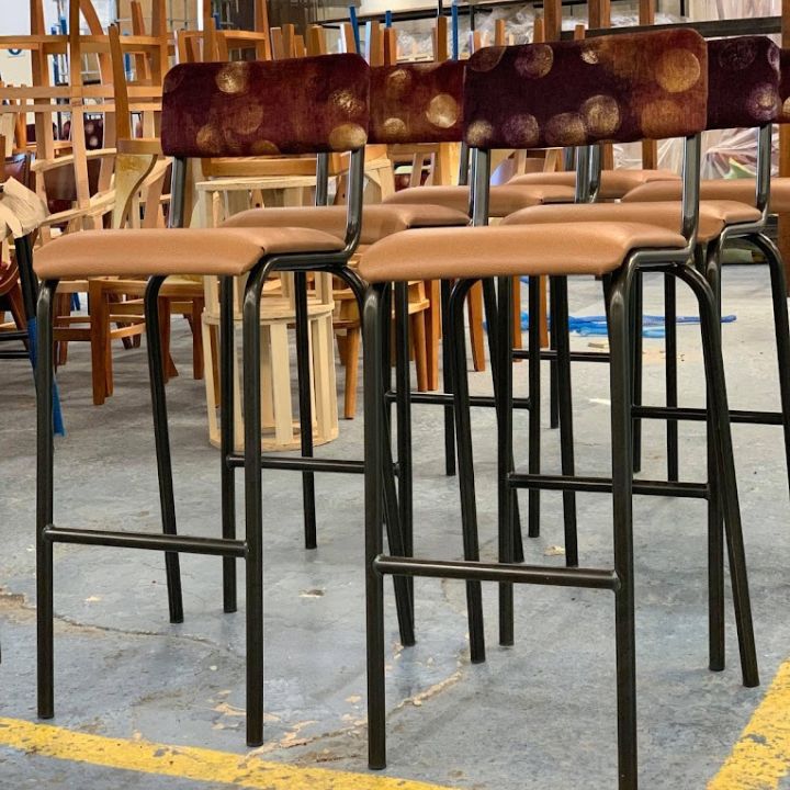 Bar stools with upholstered seats and patterned backs on black metal frames arranged in a warehouse showroom.