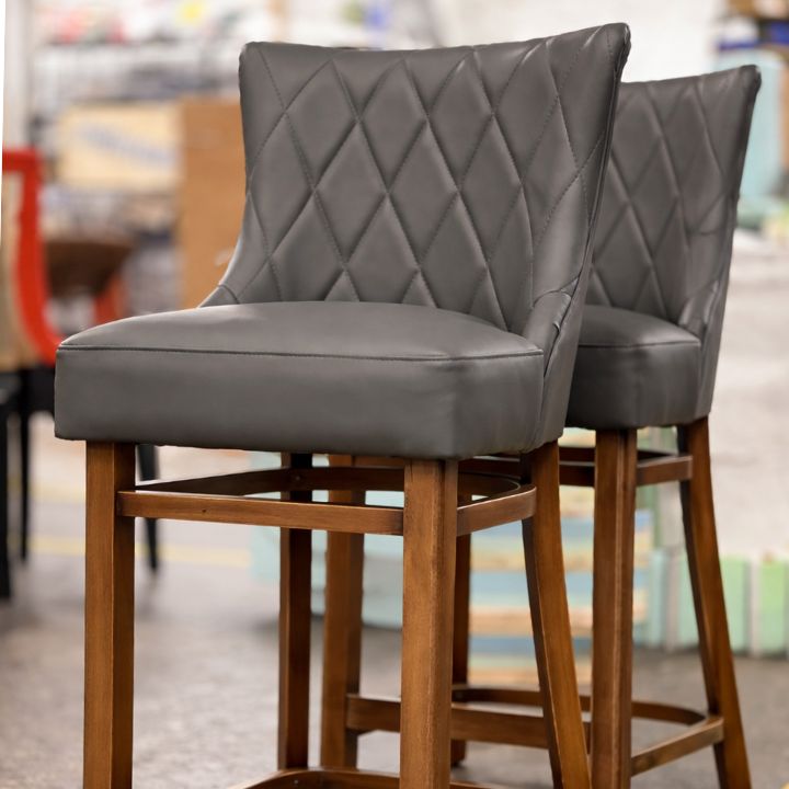 Two grey upholstered bar stools with diamond-quilted backs and wooden legs, placed in a furniture workshop with blurred equipment and materials in the background.